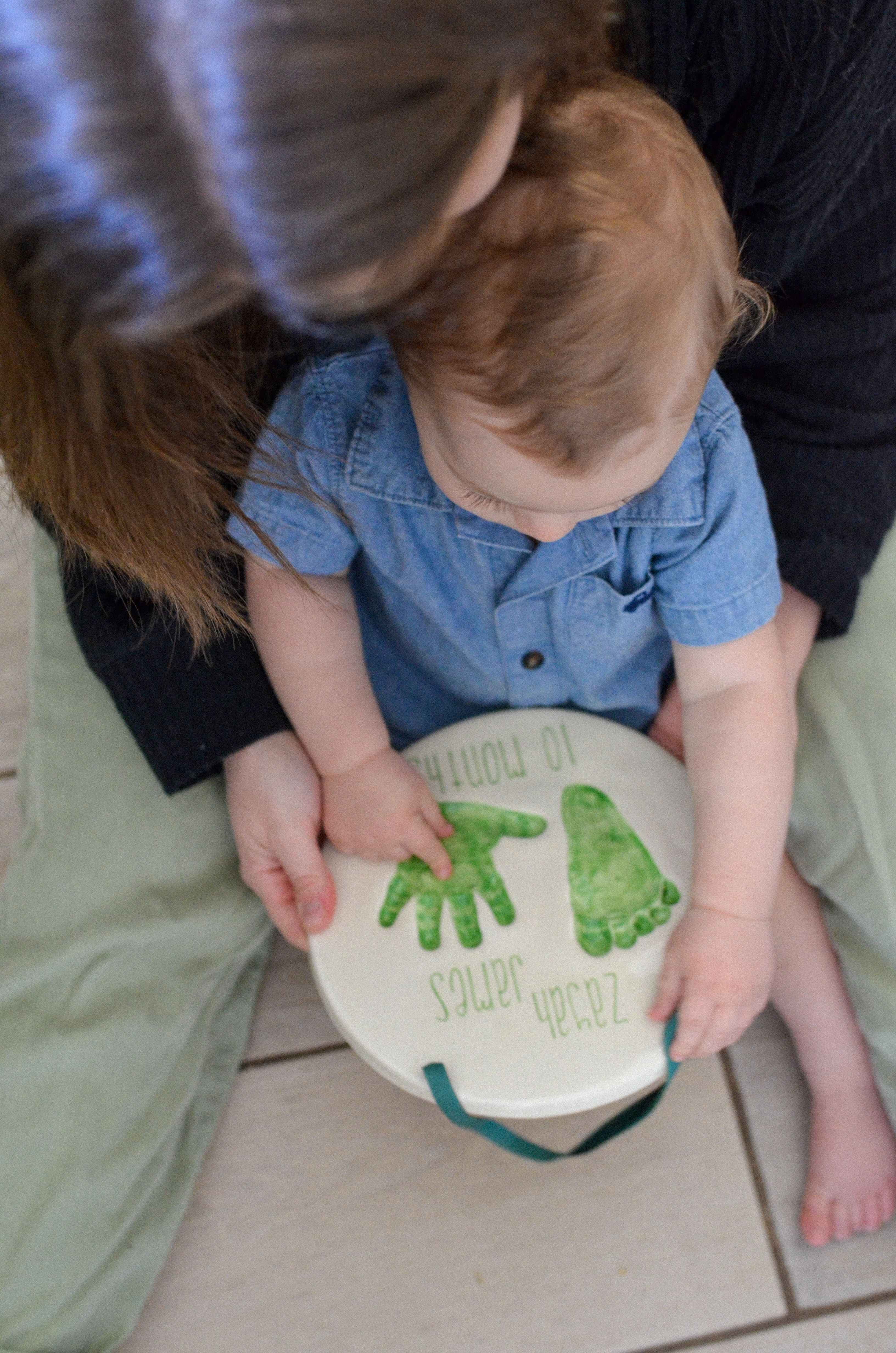 A toddler holding a finished green hand and footprint plate lettered with their name.