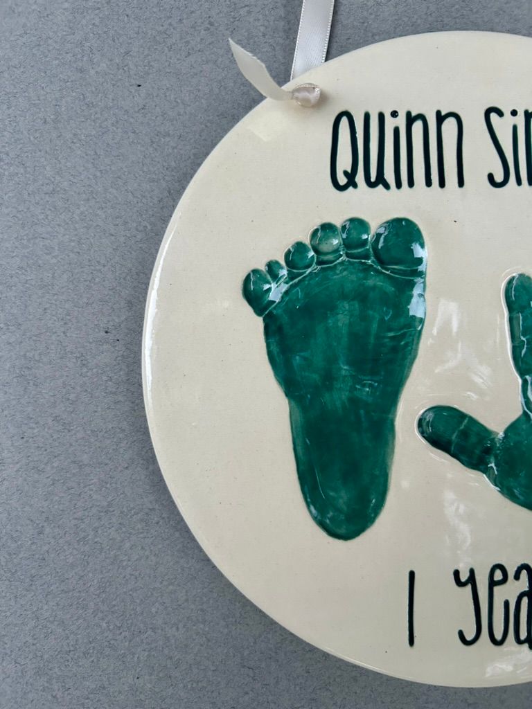 Green footprint and handprint on a white ceramic plate lettered 'Quinn Simon, 1 year.'
