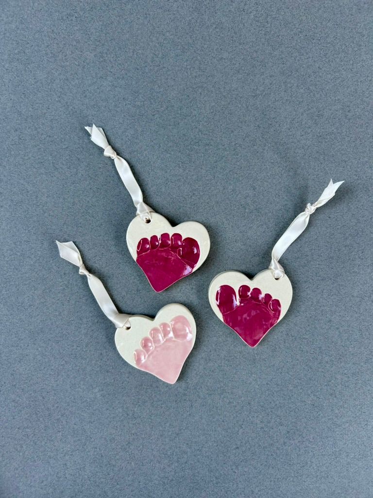 Three heart-shaped ceramic ornaments, each with a baby's toe prints in pink and red glaze.