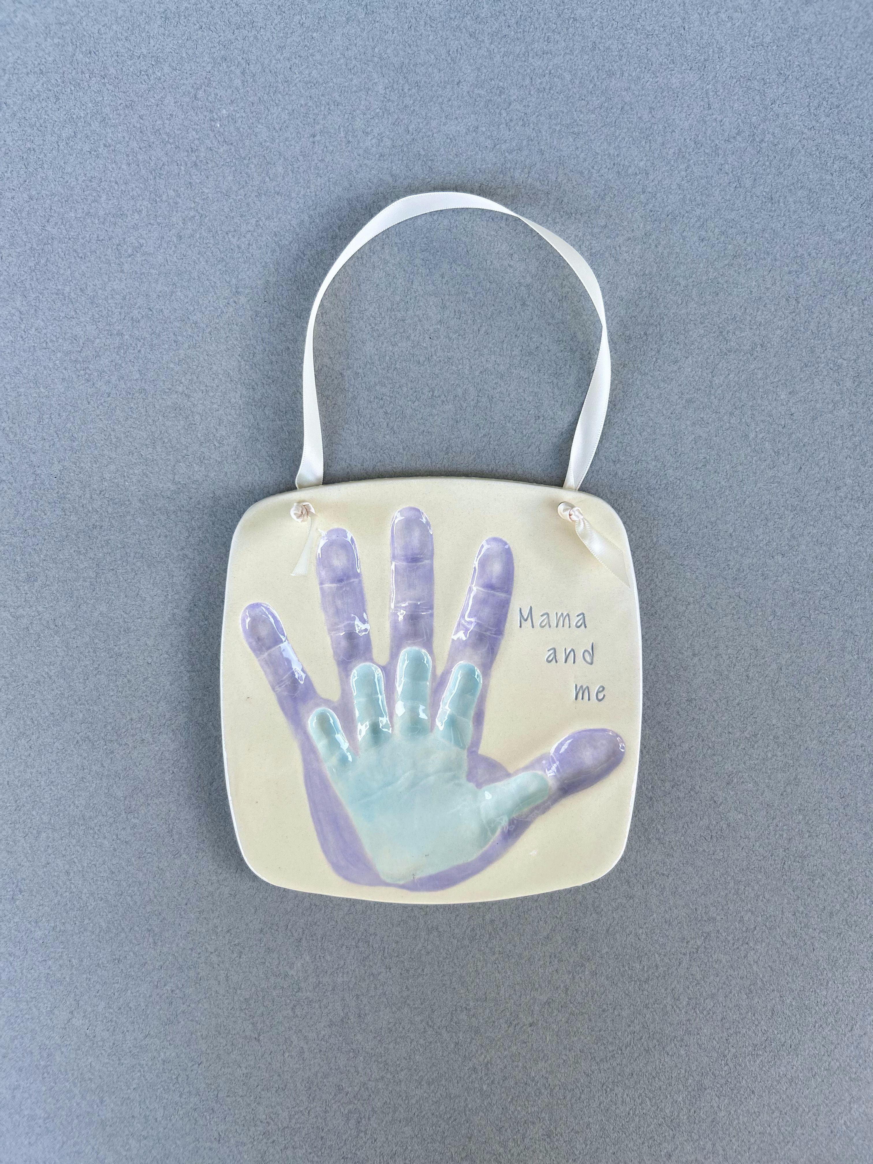 Square ceramic plaque with a mother's purple handprint and a baby's mint handprint overlapping, labeled 'Mama and me.'